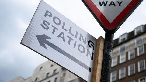 A white polling station sign with a black arrow on it attached to a sign post. Buildings can be seen towering above the sign from a distance away.