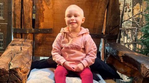 A little girl wearing a pink jumper and leggings smiling and sat on a giant wooden chair