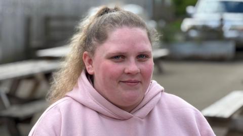 A young woman smiling at the camera. She has her hair in a ponytail and is wearing a pink hoodie. She's sitting on a picnic bench area at Forge Mill Farm in Sandwell. 