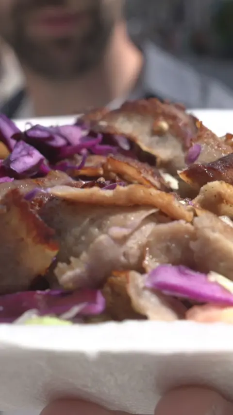A man who is out of focus holding a kebab with brown meat, green lettuce and red (purple) cabbage. The packaging is white.

