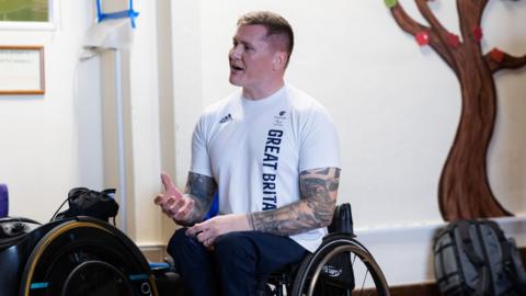 David Weir in a wheelchair speaking. A handmade tree is behind him on the wall.