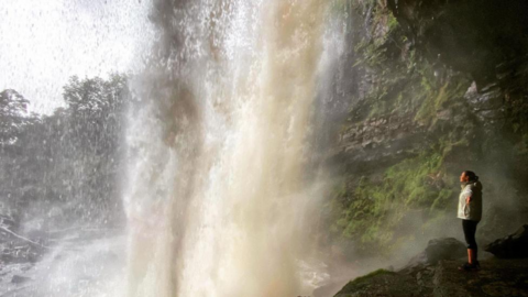 A woman stands behind a waterfall as it plunges from ground above