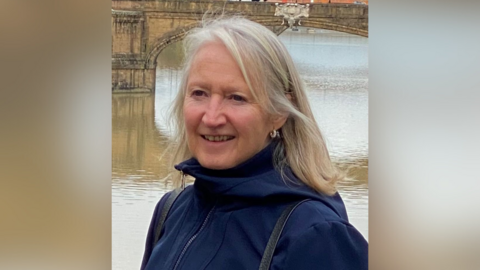 A woman with blonde hair is wearing a navy blue coat and standing in front of a river and a bridge.