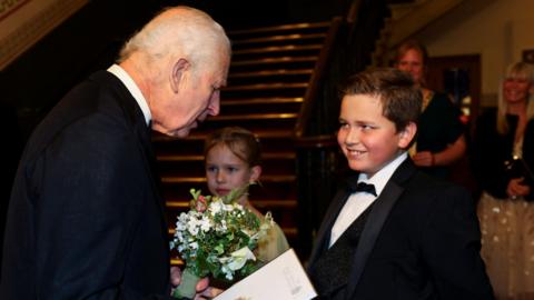 The King (L) holding a posy of flowers given to him by Iris King-Taylor (C) as he talks to Louie Cooper (R), both children of Royal Variety supporters. In the background, there is a staircase, and two women on the right.