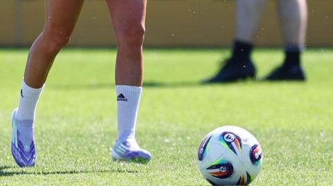 A football on a pitch with a player's legs approaching it.