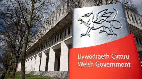 The Welsh government's Cardiff headquarters in Cathays Park, with a large silver and red sign in front of it with the words Welsh government in Welsh and English.  The grey concrete building has columns wrapping around the ground floor and two horizontal rows of windows above it. 