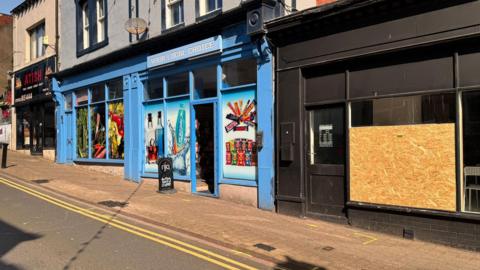 A row of shops on Senhouse Street, Maryport, where the van hit the girls. The shop on the far right has had its window boarded up. It has opening times written on the glass door and a white seat can be seen through the window. The shop looks closed. To the left there is a blue off-licence with an open door. The windows are covered up in photos of crisps, drinks and chocolate bars. There is a takeaway shop on the far right.