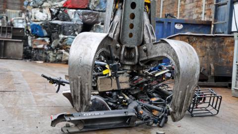 A large silver claw is pushing down on a pile of black escooters, with 'Carrera' written in white italics on, in a junkyard. There are piles of squashed metal cubes in the background and metal skips.