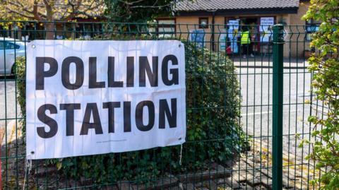 A black and white polling station poster is on a fence outside a school. People are queuing up to go in and vote.