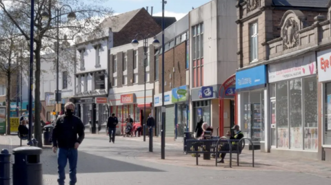 Pedestrians can be seen walking along Bilston high street. There are shops on either side, most look open, with some people sat on benches in front of the stores. The sun is shining.