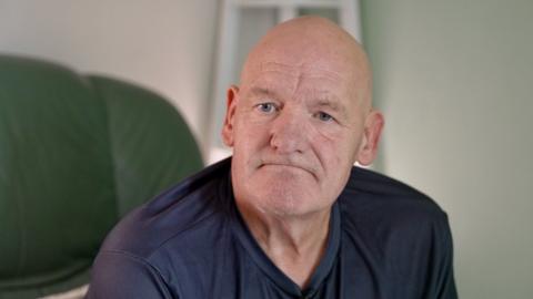 Patrick Moore sitting on a green chair looking at the camera wearing a navy blue top. He has a bald head and is looking glum at the camera.