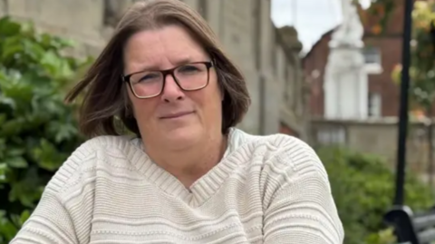 A woman with long brown hair, glasses and a white jumper with bushes behind her