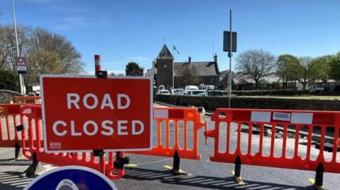 Roadworks visible in a road near St John's church. There are large red barriers and a red road closed sign. Part of the road has been dug up. 