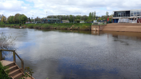 A river with a set of wooden steps in the bottom left corner. On the opposite bank there is a set of steps leading up to a low building with scaffolding outside. Other shed-like buildings are visible in the distance, further along the river.