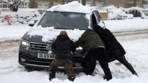 heavy snow on the ground and road with three people helping to push a car that has clearly got stuck