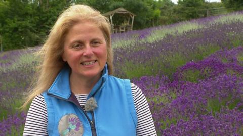 Tina Bessell stood on the left wearing a black and white striped top with a blue gilet over the top. She is smiling and has blonde hair. Behind her is a field filled with purple lavender