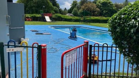 A blue paddling pool in the middle of the park with trees and a red entrance gate 