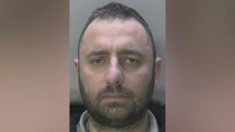 A custody image of a man with short black hair and black facial hair. He is looking into the camera with a blank expression