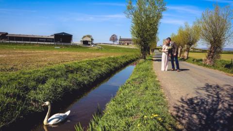 A picture with a bride wearing a wedding dress in the background kissing a groom in a suit. She is holding a bouquet and they are standing in the sunshine on a lane next to a field, with a stream running alongside them. There is a swan on the stream and farm buildings in the background.
