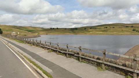 A wooden fence runs a long a road by a reservoir basin. Low green hills can be seen dotted with trees on the other side of the reservoir. 