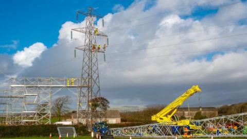 Engineers working on the replacement of an electricity pylon. The standing pylon has four workmen on its upper reaches. At its foot a plyon lies on the ground next to a yellow crane, and to the side is a high scaffolding structure with two workmen on its higher level. The scene is rural, with trees and hills in the background, and a house an what appears to be a barn just behind the scene of work. The sky has blue patches and cloud.