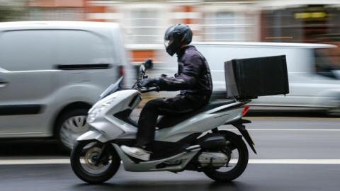 A moped driver is driving along a road. The buildings behind are blurred. The moped is silver, and the rider is dressed in black with a black helmet