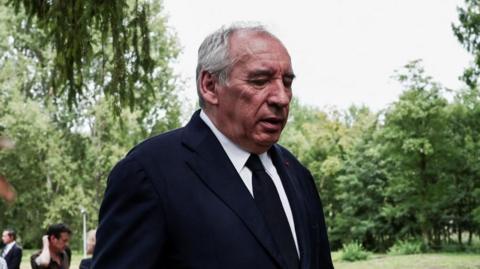 The French prime minister wears a dark jacket and a tie and with silver hair, in a garden with a backdrop of trees