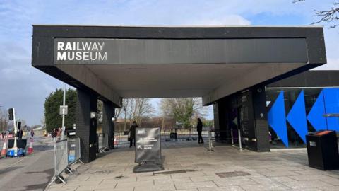 The exterior of the National Railway Museum on a sunny day. The museum building is red brick with blue and red signs and a bright yellow welcome sig above the entrance.  Two adults and a child are walking into the museum. 