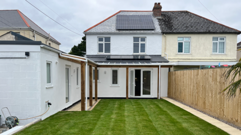 Backyard of a house with a green lawn, solar panels on the roof, a patio with wooden posts and overhang, an adjacent building on the left, and a wooden fence on the right under a cloudy sky.