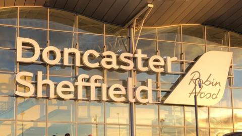 In sunset, a photo of the sign 'Doncaster Sheffield' sign on the side of the glass-paned airport building. 