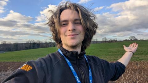 Isak Sandgren-Clarke in a selfie taken on a training walk. He is wearing a black jumper and a lanyard with 'staff' written on it. He has quite long dark blonde hair and stubble and is smiling. His arm is outstretched towards the distance.