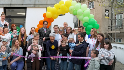 A large group of people are standing under an archway made of balloons while a woman cuts a ribbon. 