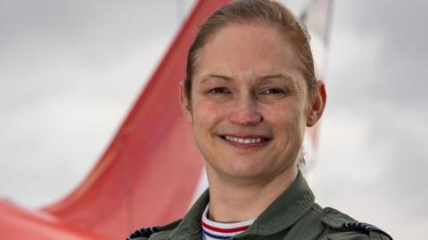 Head and shoulders shot of a smiling Wing Cdr Sasha Nash. She has fair hair which is tied back and is wearing overalls. There is the tail section of a Red Arrows jet in the background.