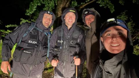 Four people dressed in waterproof black police anorak with hoods. Two have head torches and one can be seen with walking sticks. There is green vegetation behind them.