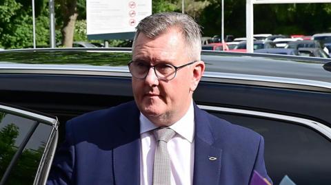 A man exiting a car - he is standing up just at the open doorway of the back seat. He has short greying hair and is wearing glasses. The top of his torso is visible, he's wearing a blue suit jacket, white shirt and tie with a black lined pattern on a white background. The car he is exiting is black. In the background the tops of other cars in a car park are visible