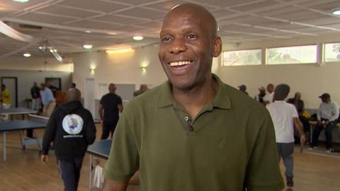 Chidi Umeano standing in a room, wearing a green top, with men playing table tennis behind him. There are windows in the room. He is smiling and looking off to the camera. 