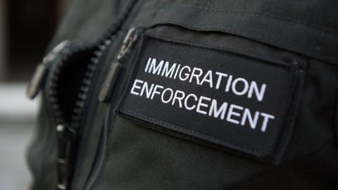 A close-up shot of a black immigration enforcement vest. The vest has 'IMMIGRATION ENFORCEMENT' written on it in white writing.
