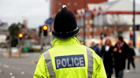 File photo of a police officer, standing with his/her back to the camera and wearing a police helmet and yellow hi-vis jacket with POLICE on the back.
He is standing next to a traffic junction in a busy town or city. Pedestrianised can be seen crossing the road behind him.