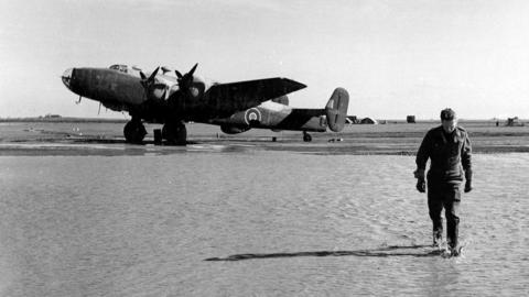 Black and white photo of an officer walking away from a plane parked on a flooded airfield