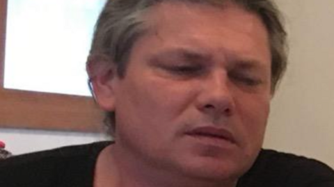 A person with short, grey-streaked hair sits indoors, wearing a black T‑shirt. They appear to be looking downward with a neutral or slightly pensive expression. A wooden doorframe and light-colored walls are visible in the background.