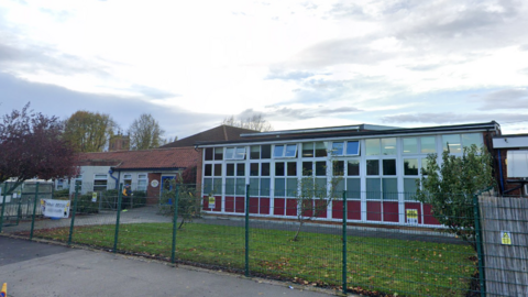 A single-storey building with brick walls, large glass windows, and a red-tiled roof, enclosed by a green metal fence. Trees, grass, and fallen leaves are in the foreground under a partly cloudy sky.
