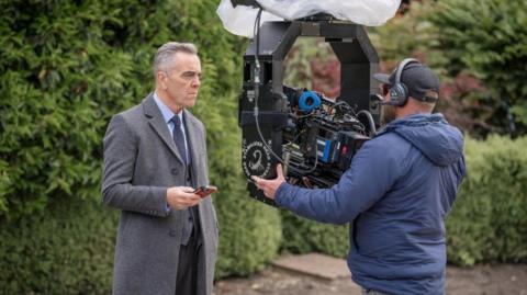 James Nesbitt, who is wearing a grey coat with suit and tie, is standing in front of a man who is holding a camera in a park during filming. 