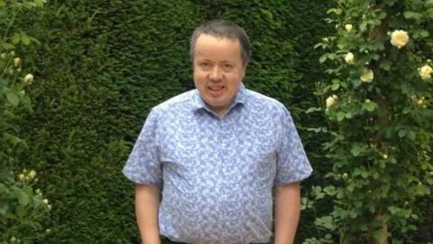 A man with grey hair smiling at the camera i front of green bushes. He is wearing a short sleeved blue patterned shirt.