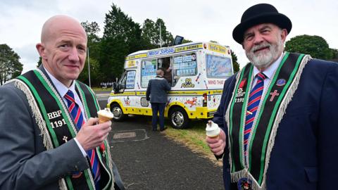 Two men with Mr Whippy ice creams standing in front of an ice cream van. They are wearing dark suits and black sashes with green trim, the man on the right has a black bowler on and the man on the left is bald.