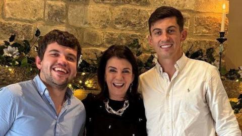Harvey Isaac, Dawn Harper and Charlie Isaac are huddled together all looking at the camera with big smiles. They are pictured in front of a large Cotswold stone fireplace and chimney breast. The brothers both have dark short hair and are wearing button up shirts. Dawn is wearing a chunky silver necklace over a black embellished dress. The mantle piece is decorated with fairy lights, candles and festive foliage.