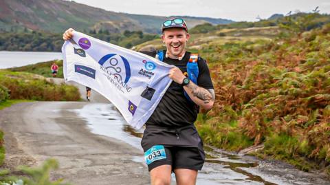 The image shows a man running with a banner in his hands. Behind him marshes can be seen and he is smiling at the camera.