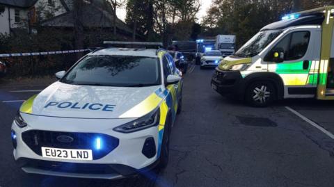 Two white police cars and an ambulance parked on a road with blue lights on. Blue and white police tape surrounds the vehicles at the scene. In the background vehicles can be seen queuing on what looks like a residential street, with trees both sides of the road and a house to the left.