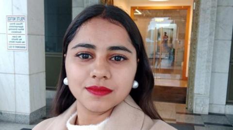 Harshita Brella looks into the camera for a selfie while standing outside a building. Her hair is open, and she is wearing bright red lipstick and pearl drop earrings.