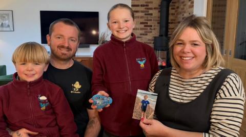 A man, woman, boy and girl smiling at the camera in a living room. The boy, on the far left, has short blonde hair and is wearing a burgundy fleece jacket. Behind him is a man with short brown hair who is wearing a black top. Next to him is a girl, who is standing up and wearing a burgundy fleece jacket. She is holding up a painted rock which has the words Billy's Boulders printed on it in rainbow letters. To her right is a woman who has chin-length blonde hair and is wearing a stripey long-sleeved t-shirt under dark-grey dungarees. She is holding up a photo of a boy wearing a blue football kit on a beach.