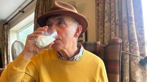 Frank in an armchair in his living room drinking a glass of mare's milk. He is wearing a yellow jumper and a tan coloured wide brimmed hat.